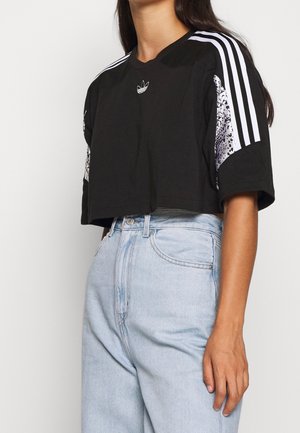 Kvinde iført en sort, kort Adidas T-shirt med hvide striber på ærmerne og lyseblå højttaljede jeans.