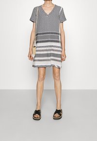 Czarna i biała sukienka w wzory, krótka, z dekoltem w serek, krótkimi rękawami i różnorodnymi paskami. Założona z czarnymi sandałami na krzyż i małą torebką.