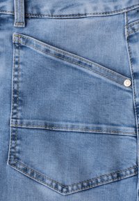 Poche de jean en denim bleu clair avec une couture orange visible et un petit rivet métallique sur le côté droit près du bord supérieur.