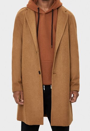 Mand iført en kamelbrun uldfrakke over en rustfarvet hættetrøje med sorte snore og sorte bukser, vist fra halsen til midt på låret.