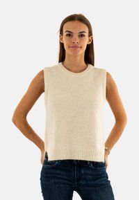 Maglione senza maniche di colore crema lavorato a maglia con scollatura e orlo a coste. Tessuto morbido, taglio dritto e spacchi laterali. Abbinato a jeans blu scuro.