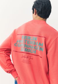 Felpa color corallo con testo stampato di nomi di città in contorni blu e rossi, scollo tondo e polsini a costine. Tessuto morbido al tatto.