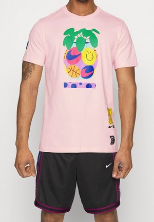 Mężczyzna ubrany w różowy t-shirt z kolorowym wzorem ananasa oraz czarne spodenki Nike z fioletowymi paskami, stojący na białym tle.