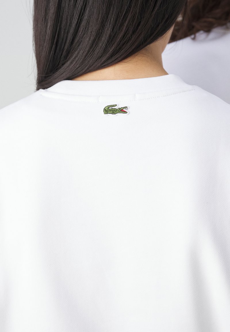 T-shirt bianco in cotone con colletto a girocollo e un piccolo logo di coccodrillo verde ricamato sul retro, vicino al colletto.