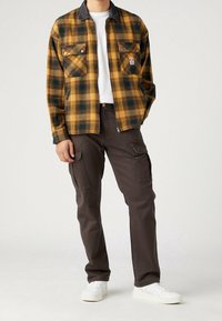 Mies, joka on pukeutunut kelta-mustaan ruutujakkuun valkoisen paidan päällä, tummanruskeisiin cargo-housuihin ja valkoisiin lenkkareihin, seisoo yksi käsi taskussa.