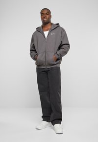 Grå hettejakke med glidelås og kengurulomme, ribbede ermer og kant; brukt over en hvit skjorte, kombinert med svarte denimjeans og hvite sneakers.