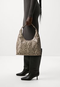 Sac à main hobo en cuir imprimé serpentin beige avec une silhouette courbée et une quincaillerie dorée, tenu par une personne portant une robe grise côtelée et des bottes noires.