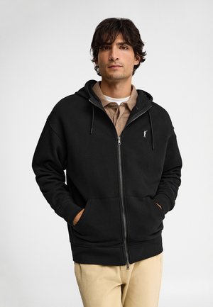 Hombre con cabello oscuro, vestido con una sudadera negra con cremallera sobre una camisa marrón y pantalones beige, de pie con las manos en los bolsillos de la sudadera.