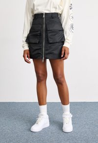 Black, zip-front mini skirt with two large, front flap pockets, made from smooth, shiny material. Worn with white sneakers and socks.