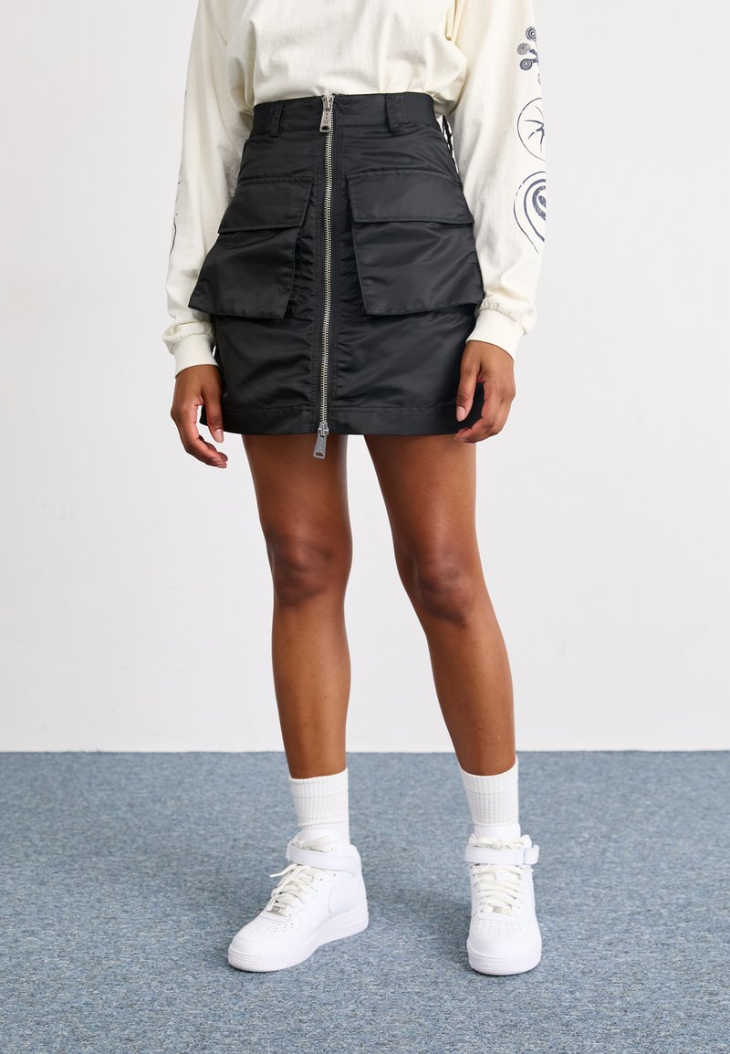Black, zip-front mini skirt with two large, front flap pockets, made from smooth, shiny material. Worn with white sneakers and socks.