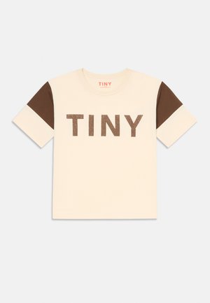 Krem t-skjorte med brune korte ermer, med stor brun glittertekst "TINY" på forsiden. Standard crew neck-design.