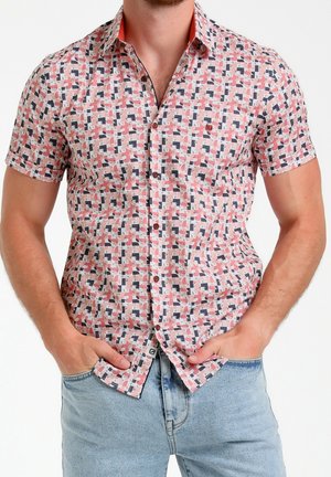 Mężczyzna ubrany w koszulę na guziki z krótkim rękawem o geometrycznym wzorze w różowym, granatowym i beżowym kolorze, ręce w kieszeniach, w połączeniu z jasnoniebieskimi dżinsami.