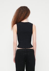  Γυναίκα με μακριά, σγουρά μαλλιά, φορώντας αμάνικη μαύρη υφασμάτινη μπλούζα και μαύρα τζιν, εμφανιζόμενη από πίσω πάνω σε απλό λευκό φόντο.