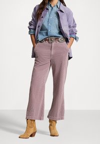 Jasnofioletowa kurtka z sztruksu nałożona na niebieską koszulę dżinsową, zestawiona z szerokimi spodniami w kolorze mauve i beżowymi kowbojkami, ozdobiona srebrną klamrą pasa.