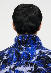 Άτομο με κοντά μαύρα μαλλιά, που φαίνεται από πίσω, φοράει fleece μπουφάν με ψηλό γιακά και αφηρημένο σχέδιο σε μπλε, μαύρο και άσπρο.