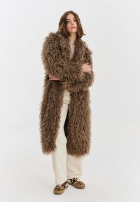 Dlouhý, huňatý kabát z hnědého umělého kožíšku, otevřený přední střih, v kombinaci s světlými kalhotami a vzorovanými hnědými botami. Měkká textura, výrazný vzhled.