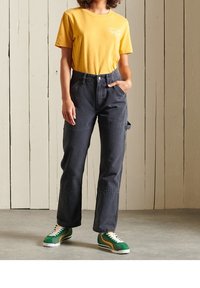 Camiseta de algodón amarilla con logo, combinada con pantalones vaqueros rectos de color gris oscuro y zapatillas verdes con acentos amarillos. Un conjunto casual sencillo.