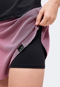 Main soulevant le bord d'une jupe rose et noire à motifs, révélant un short de cyclisme noir avec une étiquette "ENDLESS" sur la cuisse.