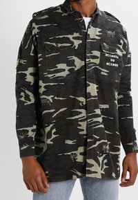 Camicia con motivo mimetico in verde scuro e nero, con due tasche sul petto e la scritta "SEASON NO SEASON", maniche lunghe e vestibilità comoda.