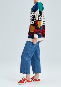 Pull en tricot avec un design audacieux en patchwork multicolore comportant des motifs floraux et géométriques ; associé à un jean en denim ample et des baskets rouges.