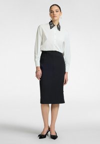 Camicia bianca con colletto nero decorato, abbinata a una gonna a tubino nera con bottoni sul lato e tacchi neri con dettagli a fiocco.
