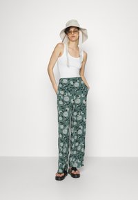 Pantalonas largas de cintura alta com padrão floral verde, combinadas com uma t-shirt branca, um chapéu tipo balde de cor clara e sandálias pretas.