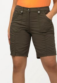 Brown knee-length shorts with multiple pockets, button closure, worn by a person with one hand in pocket, set against a plain white background.