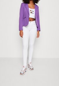 Fialový blazer přes bílý crop top s motivem motýla, doplněný bílými skinny džíny a vícebarevnými sportovními botami.
