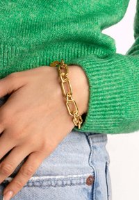 Bracciale in oro con maglie allungate e texture, indossato su una mano, abbinato a un maglione verde a maglia. Il tessuto in denim visibile è di un azzurro chiaro.