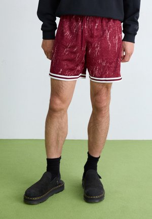 Shorts à motifs rouges en tissu texturé, avec des bandes latérales blanches et une taille élastique. Associés à des chaussures noires à enfiler et des chaussettes.