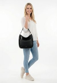 Glimlachende vrouw in wit shirt en lichtblauwe spijkerbroek houdt een zwarte leren handtas met schouderband vast, draagt beige sneakers tegen een witte achtergrond.