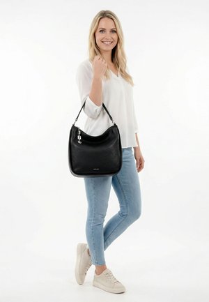 Lächelnde Frau in weißem Oberteil und hellblauen Jeans hält eine schwarze Lederhandtasche mit Schulterriemen, trägt beige Sneaker vor weißem Hintergrund.