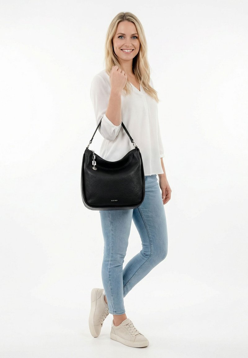 Glimlachende vrouw in wit shirt en lichtblauwe spijkerbroek houdt een zwarte leren handtas met schouderband vast, draagt beige sneakers tegen een witte achtergrond.