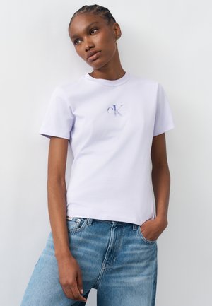 Donna con capelli intrecciati che indossa una t-shirt bianca con il logo Calvin Klein e jeans blu, in piedi con una mano in tasca davanti a uno sfondo uniforme.