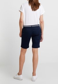 Femme portant des baskets blanches, un short bleu marine jusqu'aux genoux avec une ceinture tressée, et une chemise blanche à manches courtes, posant devant un fond uni.