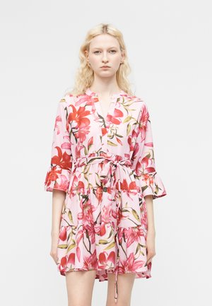 Moteris su šviesiais banguotais plaukais, dėvinti rožinę gėlėtą suknelę su trimis ketvirčiais raukiniais rankovėmis ir juosmens raišteliu, stovinti priešais vientisą foną.