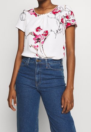 Vrouw die een witte blouse met korte mouwen met roze bloemenprint en blauwe high-waisted jeans draagt, staand tegen een effen achtergrond.