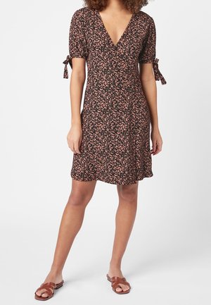 Femme portant une robe courte noire avec imprimé floral rose, col en V, manches courtes bouffantes avec nœuds noués et sandales plates marron.