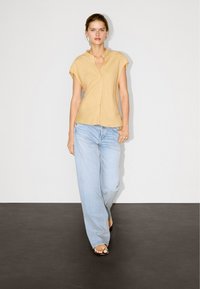 Senfgelbe kurzärmlige Knopfbluse mit Kragen, kombiniert mit hellblauen weiten Jeans und schwarzen Absatzsandalen. Neutraler Hintergrund.