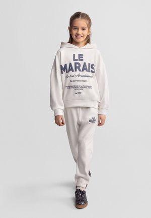 Młoda dziewczyna uśmiechająca się, ubrana w biały kaptur i spodnie dresowe z napisem "LE MARAIS", idąca naprzód na jednolitym szarym tle.