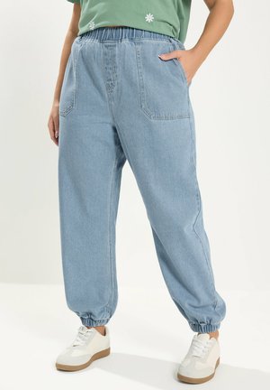 Person trägt hellblaue Jeans-Jogginghosen mit elastischem Bund und großen Fronttaschen, kombiniert mit weißen Sneakern, steht mit einer Hand in der Tasche.