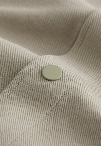 Beige textured fabric with diagonal ribs, featuring a green round button detail sewn onto the surface. Smooth texture, soft appearance.