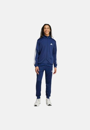 Joven de pie, vistiendo un chándal Adidas azul marino con rayas blancas y zapatillas blancas, sonriendo frente a un fondo claro.