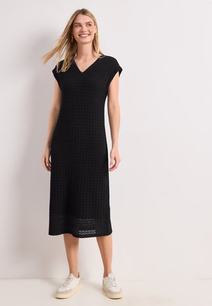 Donna sorridente che indossa un vestito midi nero lavorato a maglia senza maniche con scollo a V e scarpe da ginnastica bianche, in piedi davanti a uno sfondo chiaro e semplice.