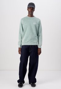 Sudadera verde claro de aspecto desgastado con puños acanalados, combinada con unos vaqueros de pierna ancha oscuros y una gorra oscura. Unos zapatos negros completan el atuendo.