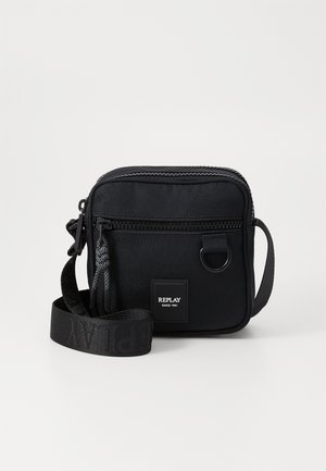 Sac bandoulière noir compact avec poche avant zippée, sangle réglable et petite étiquette logo indiquant "REPLAY SINCE 1981."