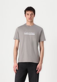 Camiseta de algodón gris con cuello redondo y mangas cortas, que presenta el logotipo blanco "Calvin Klein" en el pecho. Textura suave, corte regular.