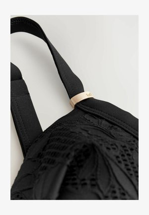 Sostén negro de encaje con tejido texturizado, tirantes ajustables y un acento de hardware en tono dorado en la tira. Patrón de encaje detallado visible.