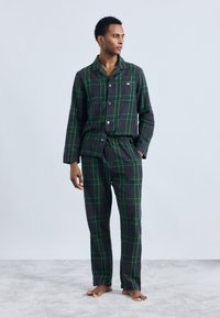 Pigiama a quadri neri e verdi realizzati in tessuto morbido. La camicia ha colletto, bottoni e maniche lunghe; i pantaloni hanno una vita elasticizzata.