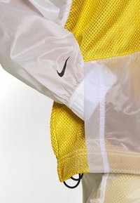 Nike vindjacka med ett genomskinligt vitt material och meshdetaljer i gult. Har elastiska manschetter och en diskret svart logotyp.